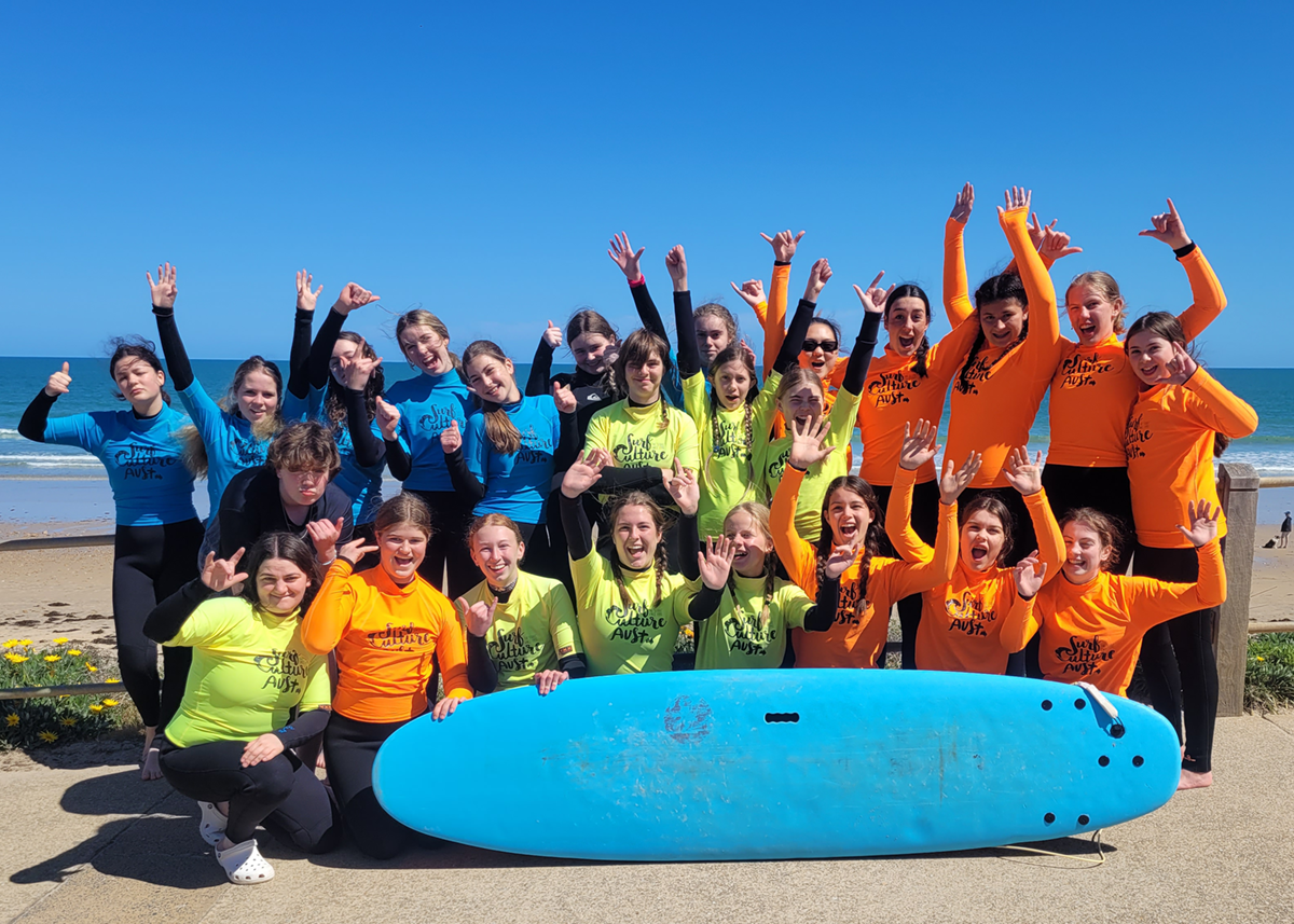 Group of Mercedes College students posing with surfboards on the beach during a surf education activity, wearing colourful Surf Culture Australia wetsuits.